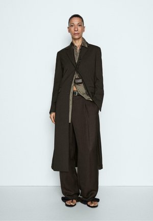 Vrouw staat en draagt een oversized donkerbruine lange jas, bijpassende wijde broek, olijfgroen overhemd, zwarte sandalen en een kleine donkere tas-ketting.