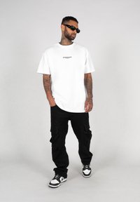 Hvit oversized t-skjorte med en liten svart logo, kombinert med svarte cargo-bukser med flere lommer, og svart-hvite joggesko.