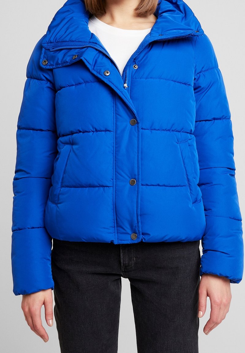 Femme portant une doudoune bleu vif avec des boutons pression sur une chemise blanche et un jean noir sur un fond clair uni.