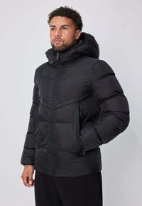 Veste matelassée noire avec capuche, dotée d'une fermeture éclair frontale, de coutures diagonales et de poches latérales, fabriquée dans un matériau lisse et léger.