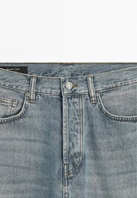 Jasnoniebieskie dżinsy z denimu o prostym kroju, z zapięciem na guziki, szlufkami na pasek oraz klasycznym designem z pięcioma kieszeniami. Brązowe przeszycia jako akcent.