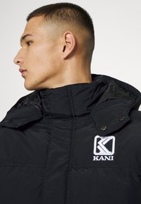 Melna pūkainā jaka ar augstu apkakli, spiedpogām un izšūtu "KANI" logo uz krūtīm. Gluda, quiltēta tekstūra.