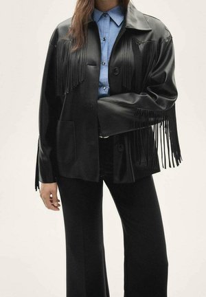 Femme portant une veste en cuir noire à franges par-dessus une chemise bleue boutonnée et un pantalon noir, sur un fond uni.