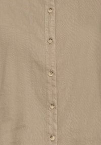 Chemise boutonnée couleur beige avec une texture douce et lisse. Six boutons assortis le long du devant, avec des coutures visibles et un détail de col.