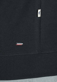 Sweat-shirt noir en coton avec un ourlet côtelé et une petite étiquette logo. Accentué de surpiqûres rouges, blanches et bleues près de l'ourlet.