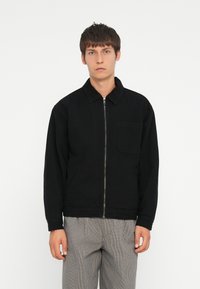 Giacca nera con zip, collo con revers, tasche frontali e maniche lunghe. Indossata con pantaloni a quadri, caratterizzati da un design semplice e informale.