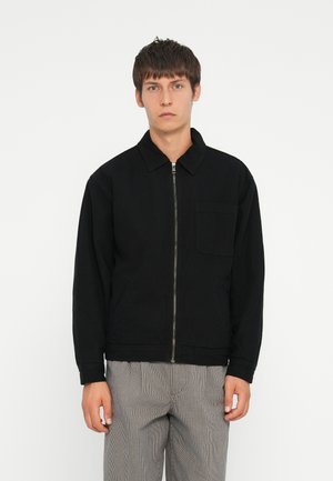 Black zip-up jacket with a collared neckline, front pockets, and long sleeves. Worn with checked pants, featuring a simple, casual design.