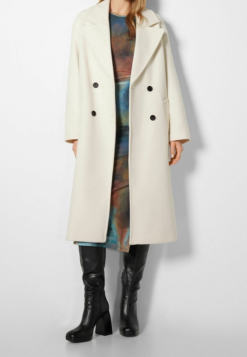 Femme portant un long manteau croisé crème sur une robe multicolore et des bottes noires à talons montantes jusqu'aux genoux, debout devant un fond uni.