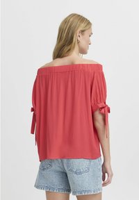 Top off-the-shoulder in tessuto corallo con finitura testurizzata, caratterizzato da maniche corte con dettagli a fiocco e una vestibilità comoda, abbinato a pantaloncini di denim.