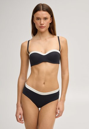 Frau steht frontal mit einem schwarzen Bikini mit weißem Rand vor einem schlichten hellgrauen Hintergrund.