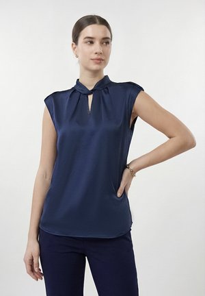 Žena v tmavomodrej bezrukávovej blúzke s výstrihom v tvare kľúčovej dierky a tmavomodrých nohaviciach stojaca s jednou rukou v bok proti bielemu pozadiu.