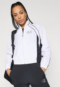 Balta un melna vēja necaurlaidīga jaka ar rāvējslēdzēju, elastīgām piedurknēm un logotipa detaļu. Apvienota ar melniem biksēm un plecu somu.