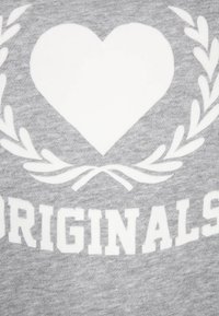 Ett vitt hjärta och en lagerkrans ovanför ordet "ORIGINALS" i fetvita bokstäver på en grå tygbakgrund.