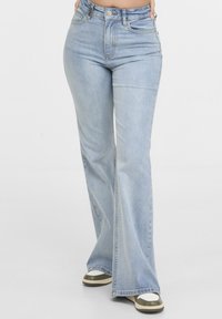 Femme portant un jean évasé taille haute bleu clair et des baskets blanches avec des accents verts, debout les mains dans les poches devant un fond uni.