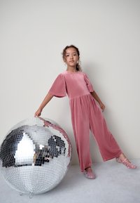 Combinaison en velours côtelé rose avec manches courtes bouffantes, associée à des chaussures pailletées, à côté d'une grande boule disco miroir. Fond simple.