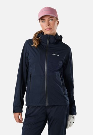 Jeune femme portant une veste zippée bleu marine Swedemount, un pantalon assorti, une casquette rose et un gant blanc à une main, tournée vers l'avant.