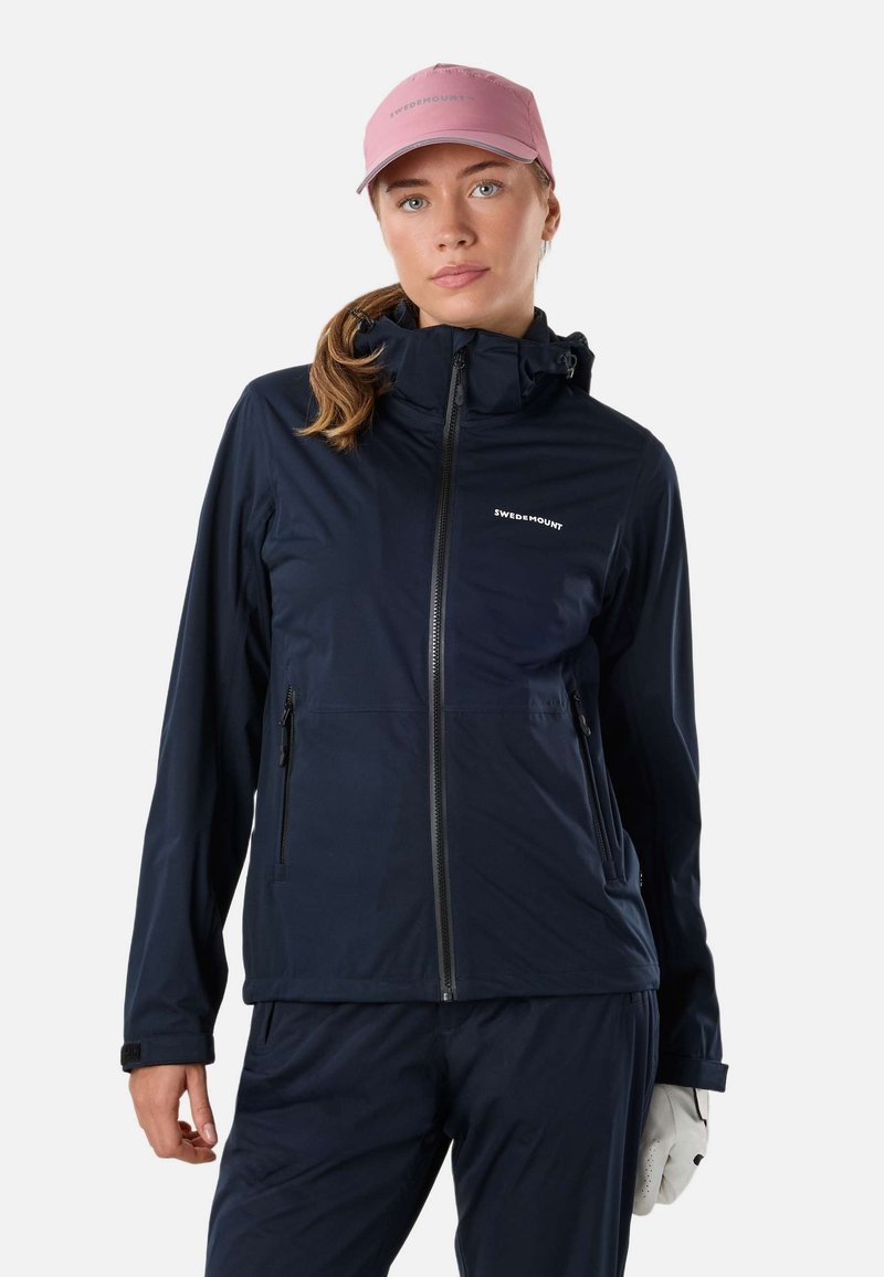 Jeune femme portant une veste zippée bleu marine Swedemount, un pantalon assorti, une casquette rose et un gant blanc à une main, tournée vers l'avant.