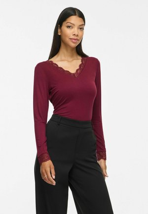 Femme portant un haut ajusté bordeaux à manches longues avec bordure en dentelle au niveau du col et des poignets, associé à un pantalon noir taille haute.