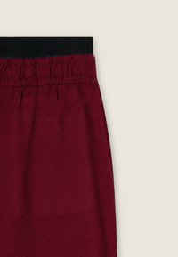 Pantalones de chándal burdeos con una cintura elástica negra que presenta un sutil logotipo. La tela tiene una textura suave y un acabado liso.