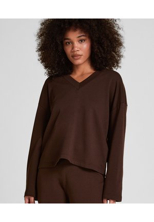 Frau mit lockigem Haar, die ein lockeres braunes Langarm-V-Ausschnitt-Oberteil und passende Hose trägt, steht vor einem schlichten grauen Hintergrund.