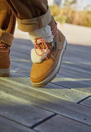 Bottes d'hiver couleur beige avec doublure en polaire blanche et lacets rouges et blancs, portées avec un pantalon en velours côtelé marron retroussé, sur une terrasse en bois.