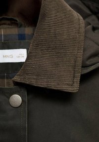 Rjav kordurojski ovratnik z teksturiranimi rebri, pripet na temno tkaninsko jakno z srebrnim pritrdilnim gumbom in karo podlogo.