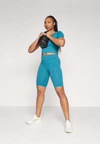 Teal activewear set featuring a fitted short-sleeve top and high-waisted bike shorts, paired with white athletic shoes and a black kettlebell.