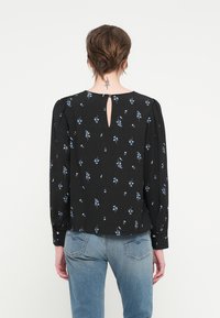 Svart bluse med små blå blomstermønstre, lange ermer og nøkkelhull detalj på ryggen. Glatt stoff med mild tekstur. Kombinert med lyse denim.