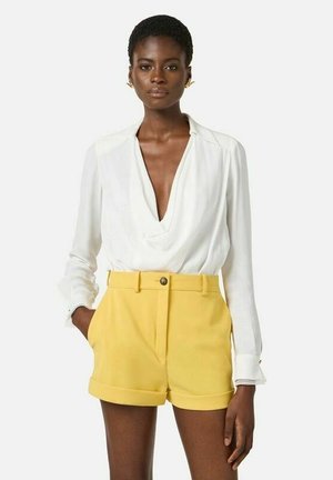 Donna che indossa una camicetta bianca a maniche lunghe con scollo profondo e pantaloncini gialli a vita alta con risvolti, in piedi con una mano in tasca.