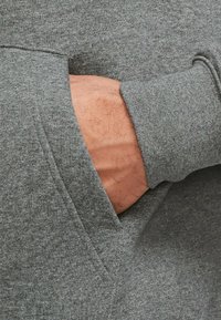 Grå sweatshirt med kangaroo-lomme. Stoffet ser blødt og struktureret ud. En hånd er indsætte i lommen, hvilket fremhæver designdetaljerne.