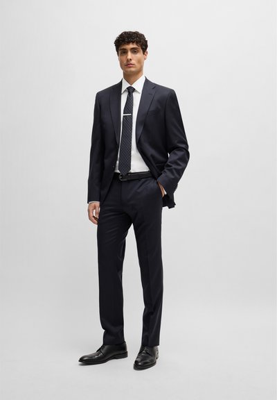 Costume marine ajusté, chemise blanche et cravate à motifs. Des chaussures noires et une pince à cravate en argent complètent le look. Tissu lisse, design sur mesure.