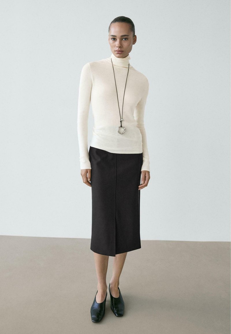 Cream turtleneck sweater, fitted; black midi skirt with a front slit. Model wears black closed-toe shoes; long necklace with a circular pendant.