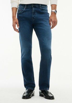 Mężczyzna w niebieskich jeansach slim-fit, czarnych mokasynach, białej koszuli z długim rękawem i srebrnym zegarku na rękę na jednolitym tle.