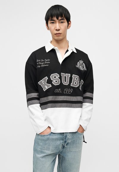 Μαύρο και άσπρο πόλο μπλουζάκι με μακριά μανίκια, κεντημένο κείμενο και λογότυπο, γκρι ρίγες και γιακά με κουμπιά. Φορεμένο με τζιν ελαφριάς πλύσης.