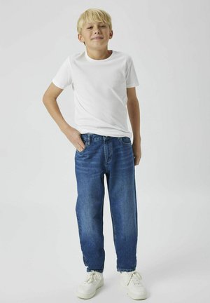 T-shirt bianca in cotone, jeans blu in denim con taglio dritto e sneakers bianche. Il tessuto è liscio; i jeans presentano una leggera scoloritura e tasche anteriori.