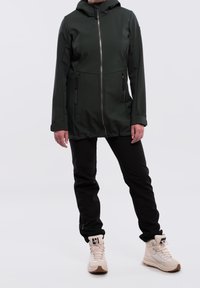 Kurtka z kapturem w ciemnozielonym kolorze, z pełnym zamkiem z przodu, dwoma bocznymi kieszeniami na zamek oraz długimi rękawami, zestawiona z czarnymi spodniami i jasnym obuwiem.