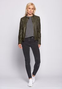 Olivgrön läderjacka med rund krage, fickor med dragkedja och figursydd design. Kombinerad med grå skjorta och svarta tajta jeans.