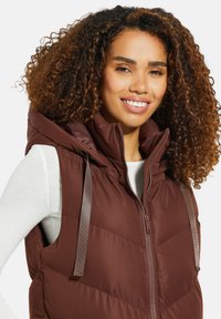 Braune, gesteppte Weste mit Kapuze, seitlichen Taschen und hellgrauen Akzenten. Das Material wirkt glatt mit einer gesteppten Textur über den Körper.