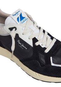 Musta ja harmaa lenkkari, jossa on mokka- ja nylonmateriaaleja, valkoiset nauhat, beige pohja ja sininen logo-osa kielekkeessä. Sisältää teksturoituja yksityiskohtia.