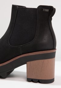 Bottine noire avec tige texturée, panneaux élastiques latéraux et talon bloc marron à finition imitation bois avec bord inférieur festonné.