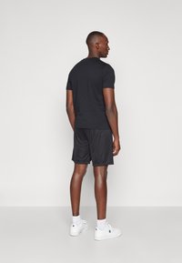 Zwarte short-sleeve t-shirt, gecombineerd met zwarte shorts. Het model draagt witte sneakers en heeft kort haar, terwijl het iets van de camera wegkijkt.