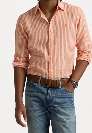 Mann trägt ein langärmeliges, orange-weiß gestreiftes Hemd mit hochgekrempelten Ärmeln, blaue Jeans, braunen Ledergürtel und Armbanduhr.