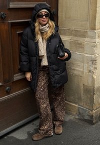 Črna puhovka s kapuco, bež pleten pulover, rjave hlače z leopardjim potiskom in rjavi semiš čevlji. V roki drži črno prešito torbico.