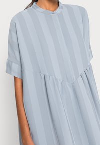 Svetle modré šaty s krátkým rukávem vyrobené z hladkého materiálu, se svislými pruhy a límečkem na knoflíky s detailem v pase. 