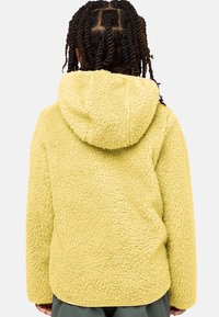 Keltainen fleece-takki, jossa on teksturoitu, pörröinen pinta ja huppu; ominaisuuksia ovat pyöreä helma ja pitkät hihat. Takakatsaus näkyvissä.