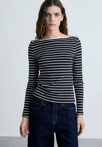 Tricou cu dungi bleumarin și crem, cu mânecă lungă și decolteu tip barcă, corp ajustat și material moale, elastic. Asortat cu pantaloni din denim închis la culoare.
