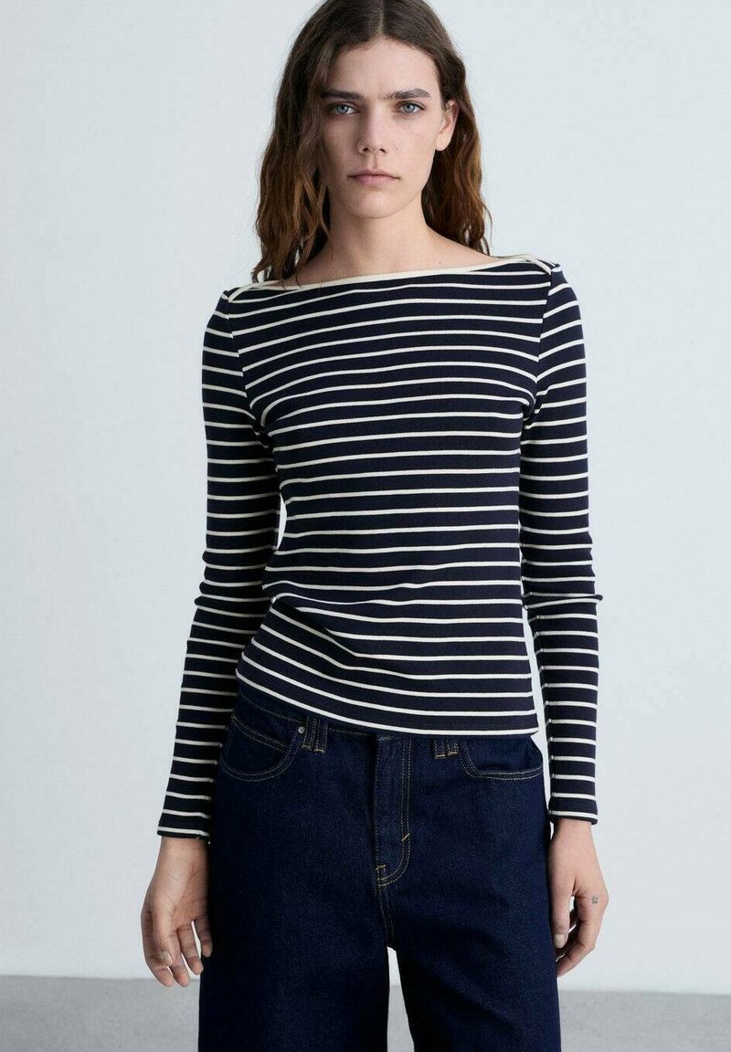 Tricou cu dungi bleumarin și crem, cu mânecă lungă și decolteu tip barcă, corp ajustat și material moale, elastic. Asortat cu pantaloni din denim închis la culoare.
