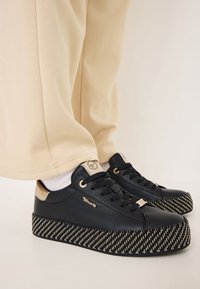 Sneaker nera con tomaia in pelle testurizzata, dettagli dorati e suola platform con un motivo a spina di pesce in nero e beige.