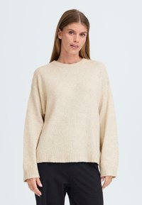 Pull en tricot beige avec une coupe décontractée, un col rond et un ourlet côtelé. Présente des manches longues et une texture douce. Associé à un pantalon noir.