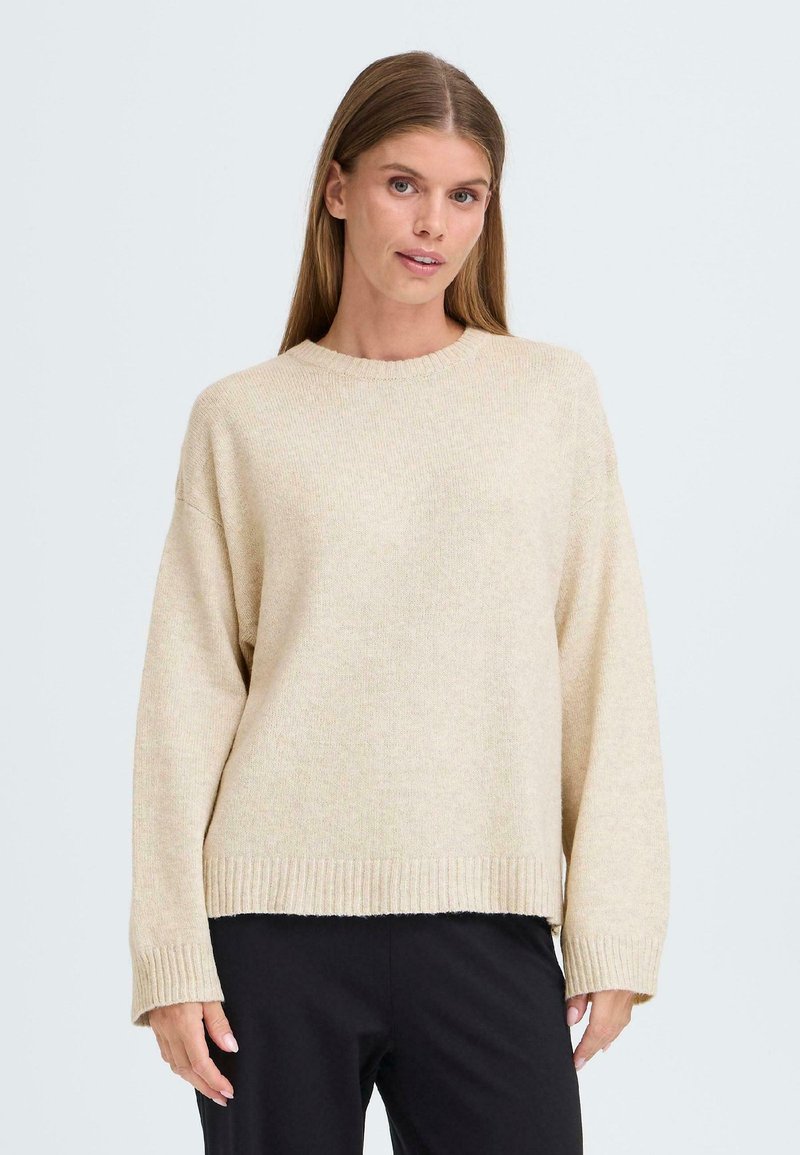 Pull en tricot beige avec une coupe décontractée, un col rond et un ourlet côtelé. Présente des manches longues et une texture douce. Associé à un pantalon noir.